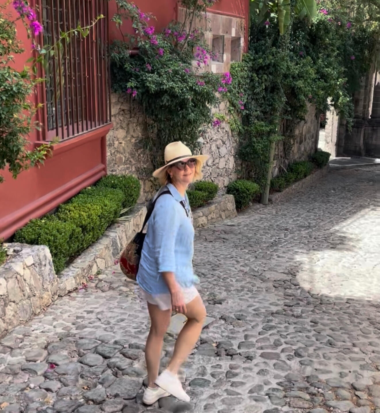 Jen Nash, founder of The Walking Coach, walking in San Miguel de Allende Mexico Photo of a woman in a hat and blue shirt with shorts walking on a cobblestone street